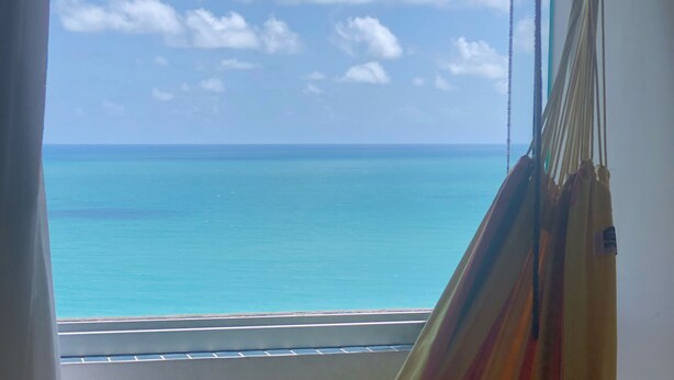 A hammock chair with a patterned pillow, overlooking a beach with clear blue water and a distant shoreline. - Flat with spectacular view of PONTA NEGRA BEACH, 55 MB Wi-Fi, garage (Natal)