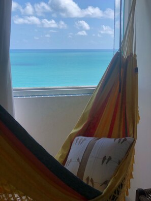 A hammock chair with a patterned pillow, overlooking a beach with clear blue water and a distant shoreline.