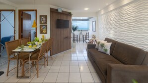 A modern living room with a brown sofa, dining area with wicker chairs, and a kitchen area with a wooden cabinet. - Flat with spectacular view of PONTA NEGRA BEACH, 55 MB Wi-Fi, garage (Natal)