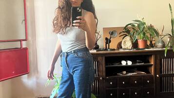 A person taking a selfie in a room with a mirror, a potted plant, and a wooden cabinet.