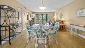 A spacious living room with a dining area, featuring wicker furniture, a ceiling fan, and a large window providing natural light.