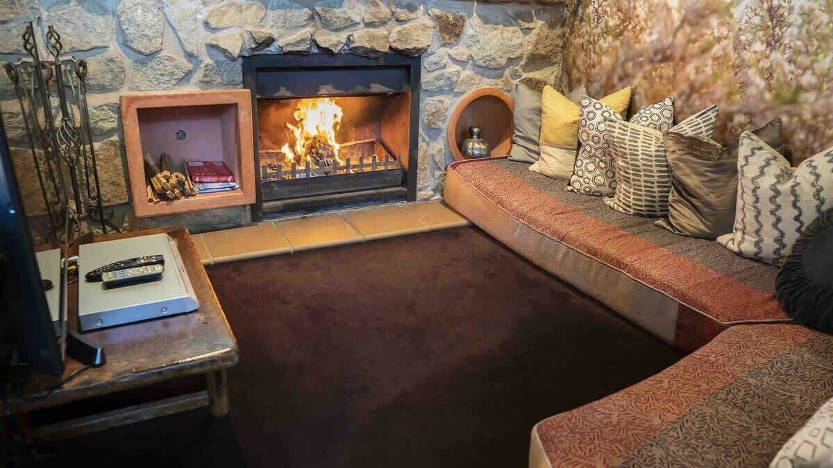 A stone fireplace with a fire, a wooden bench, a sofa with patterned cushions, and a remote control on a table.