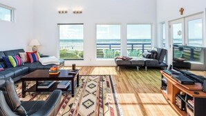 A spacious living room with a large sofa, a coffee table, and a TV stand. There are multiple windows offering a view of the ocean. - Ocean Front Beach House. Come and Enjoy The Sand, Sea, and Sun (Galveston)