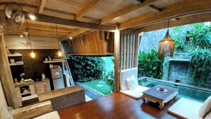 Una habitación amplia con suelo de madera, una cama, zona de estar con mesa y vistas a un jardín y piscina.