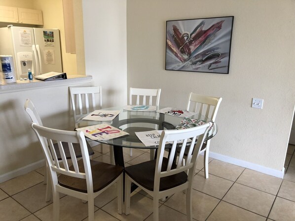 Un'area pranzo con un tavolo in vetro circondato da sedie bianche, un frigorifero con dei fogli attaccati e un quadro appeso al muro.