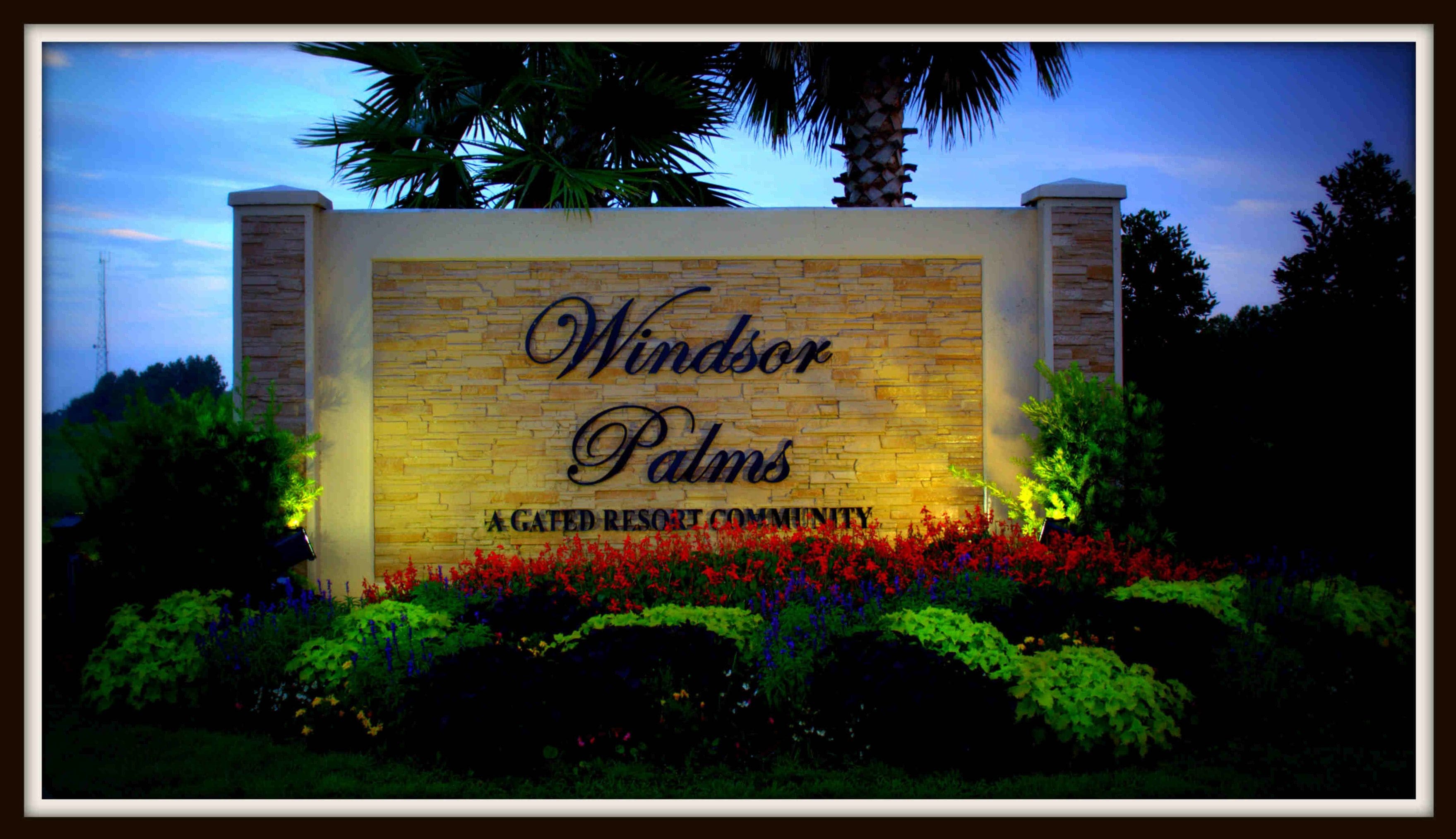 Une enseigne indiquant l’entrée de Windsor Palms, un complexe résidentiel de luxe avec portail d’accès, agrémenté d’un mur en pierre et de fleurs aux couleurs vives.