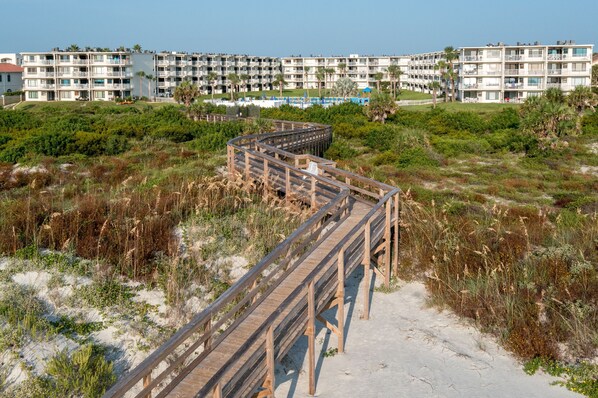 A wooden boardwalk leading to a beachfront hotel with multiple balconies and a pool area.
