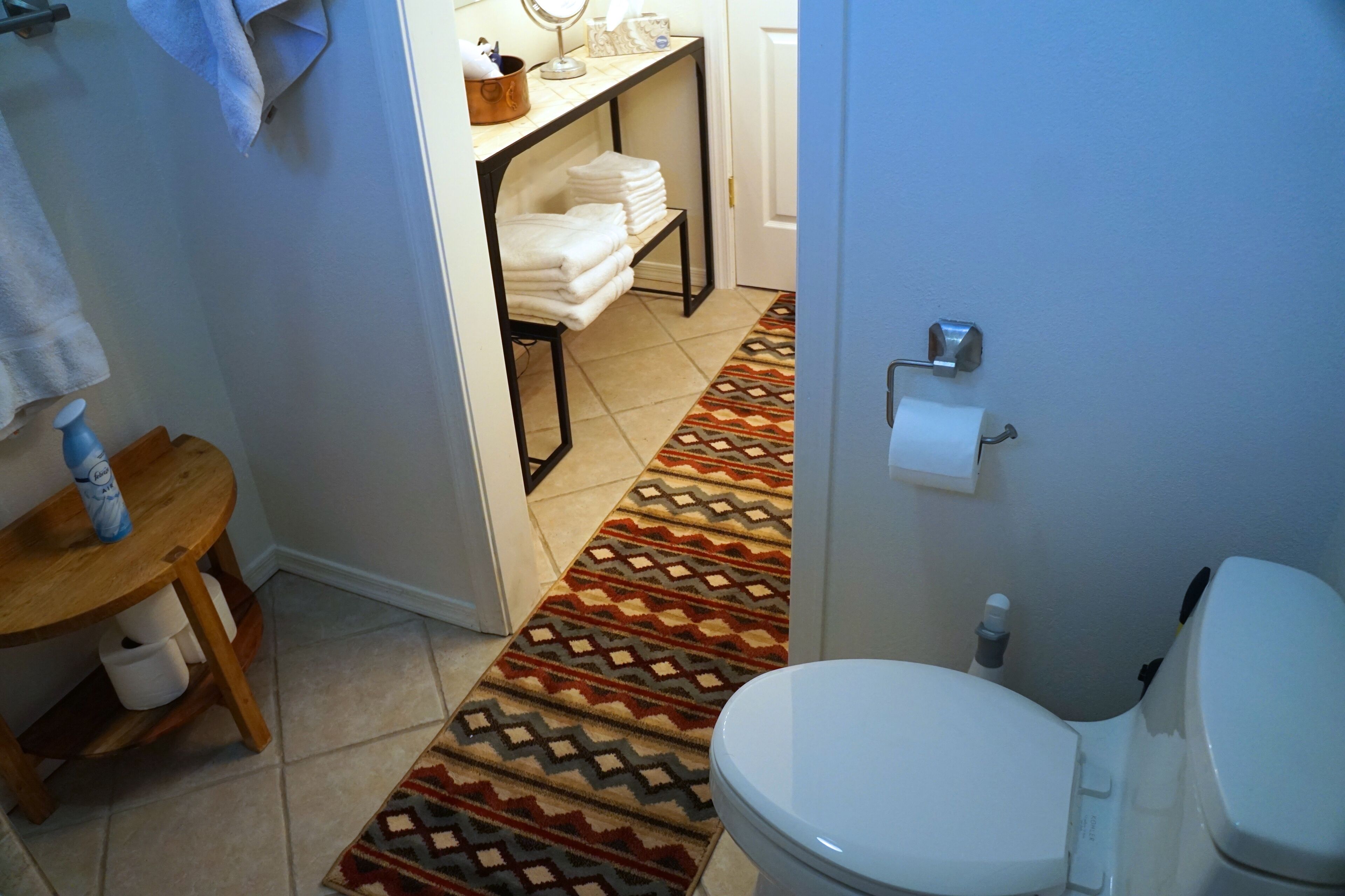 A bathroom with a patterned rug, a toilet, a sink with a mirror, and a shelf with towels.
