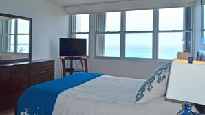 Ein Schlafzimmer mit Bett, Kommode, Fernseher auf einem Schrank und einem Fenster mit Meerblick.