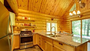 A log cabin kitchen with wooden walls, granite countertops, stainless steel appliances, and a large window overlooking the forest. - Secluded Log Cabin in NW Michigan: Hot Tub & Deck (Hersey)