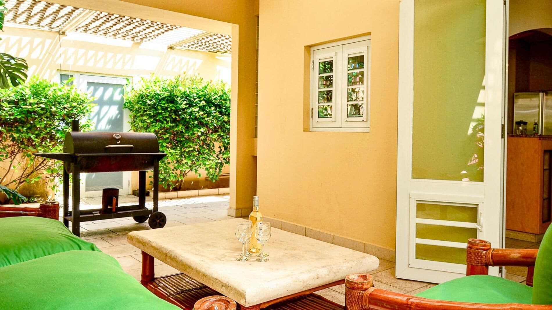 A patio with a barbecue grill, a table with glasses, and green cushions.