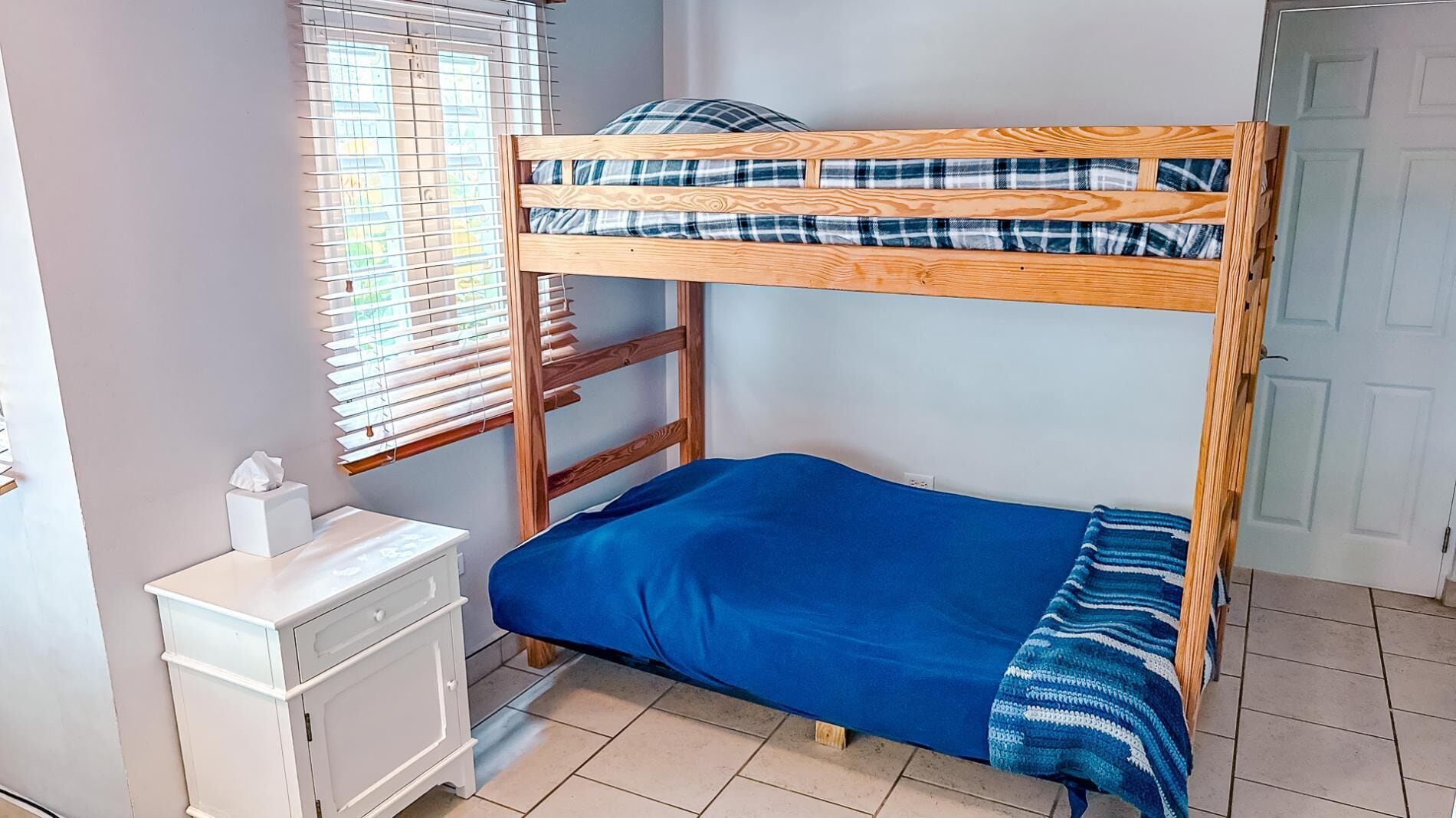 A bunk bed with a blue blanket and plaid pillow, a white bedside table with a tissue box, and a window with blinds.