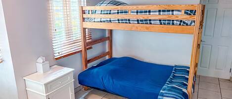 A bunk bed with a blue blanket and plaid pillow, a white bedside table with a tissue box, and a window with blinds.