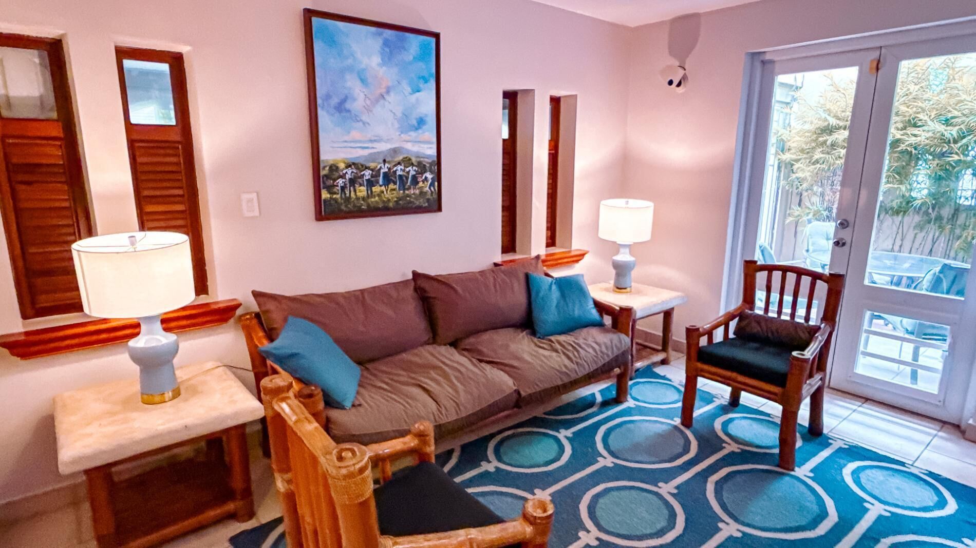 A living room with a sofa, a chair, a small table, and a lamp. There is a painting on the wall and a door leading outside.