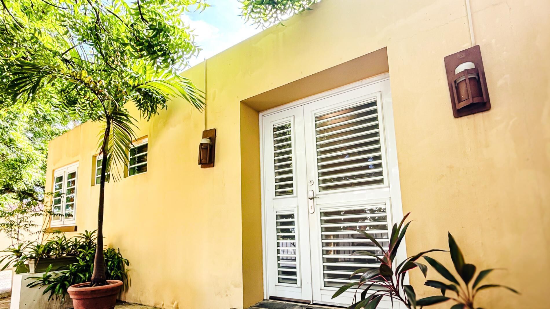A yellow building with a white door and window shutters, surrounded by potted plants.