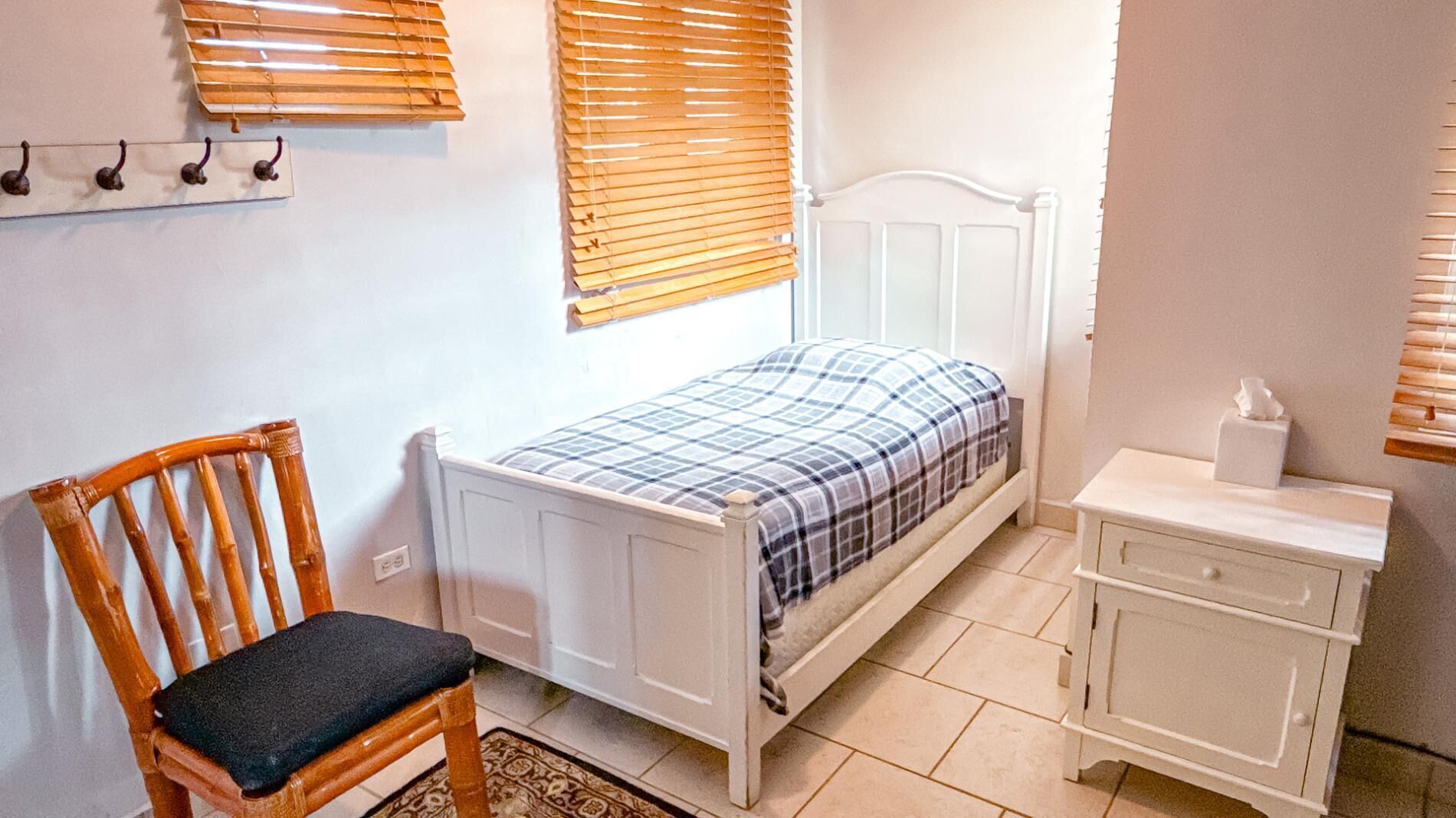Un dormitorio con una cama individual, una silla de madera, una mesita de noche con una caja de pañuelos y una ventana con persianas.
