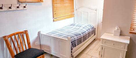 Un dormitorio con una cama individual, una silla de madera, una mesita de noche con una caja de pañuelos y una ventana con persianas.