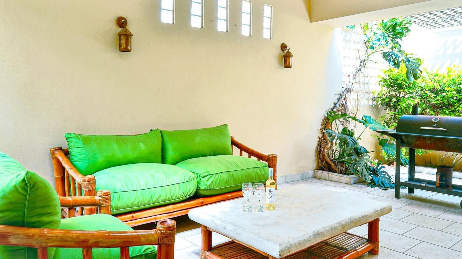 Une pièce avec un canapé vert rembourré, une petite table avec des verres et une bouteille, et un barbecue à l’extérieur.