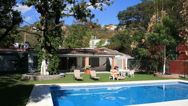 A backyard with a swimming pool, a covered patio, and a lawn with chairs and a table.