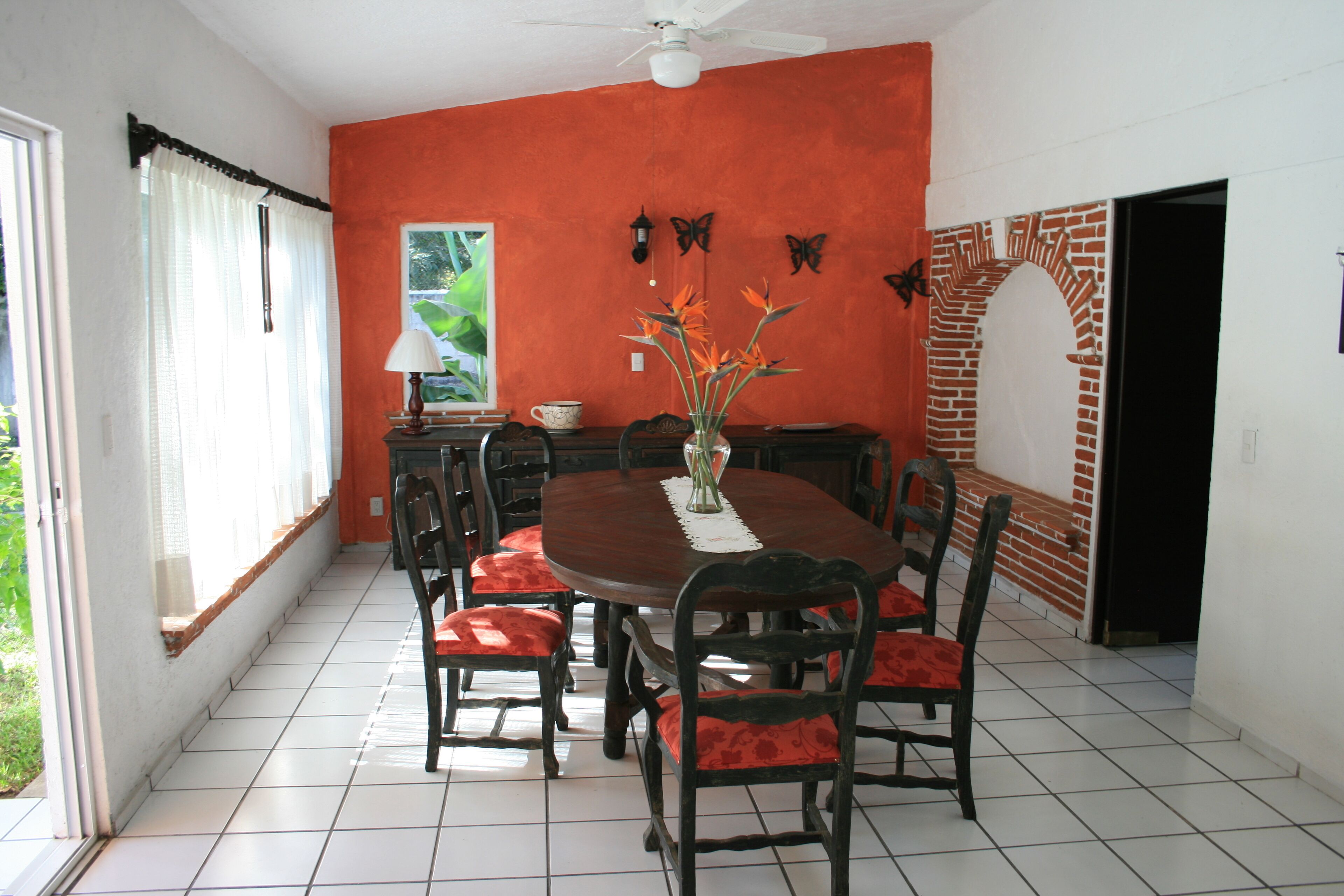 Un comedor con una mesa redonda, sillas tapizadas en rojo, un florero con flores y decoraciones de mariposas en la pared.