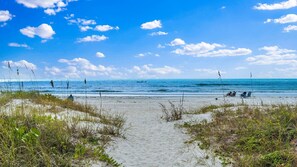 A beach with a sandy path leading to the water, some vegetation, and a clear sky.