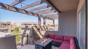 Un balcon extérieur doté d’une cheminée en pierre, d’une table à plateau en verre et d’un canapé rouge avec des coussins.