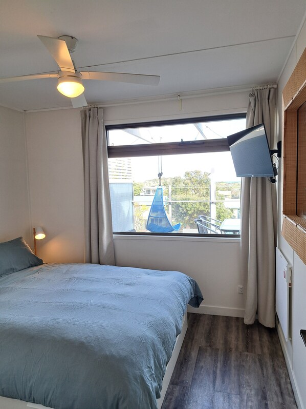 A bedroom with a bed, a ceiling fan, a window with curtains, a desk lamp, and a view of the outside.