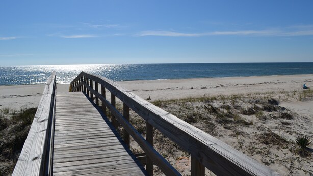 Ein hölzerner Steg führt zu einem Strand bei blauem Himmel.