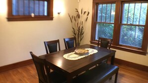 Een eetruimte met een houten tafel, stoelen, een bank, een vaas met planten en ramen met uitzicht op groen.