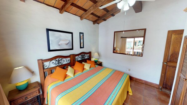 A bedroom with a wooden ceiling, a bed with colorful bedding, a nightstand with a lamp, and a window with a view of the outside.