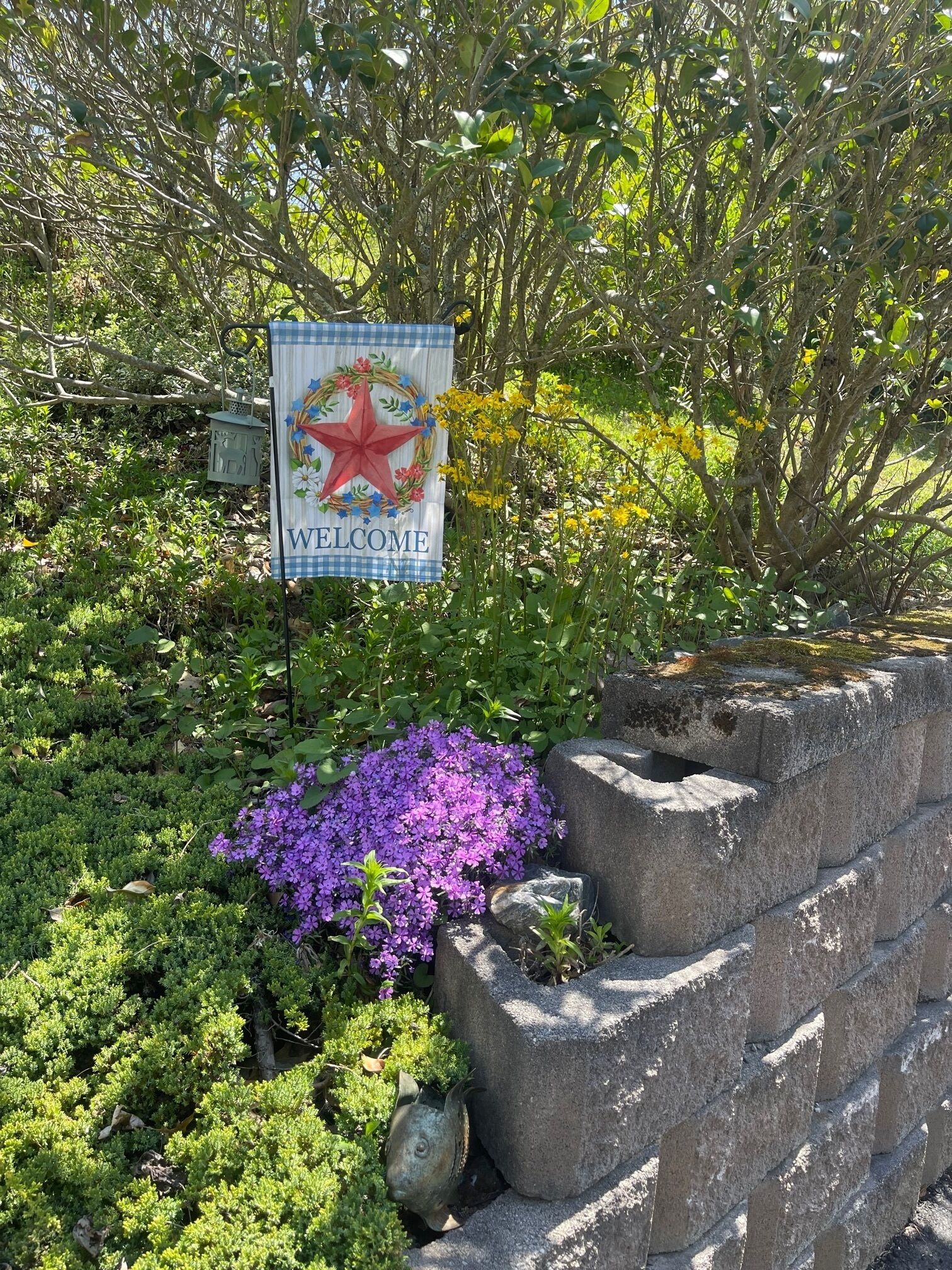 A welcome sign with a star and floral wreath, surrounded by lush greenery and a stone wall.