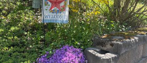 A welcome sign with a star and floral wreath, surrounded by lush greenery and a stone wall.