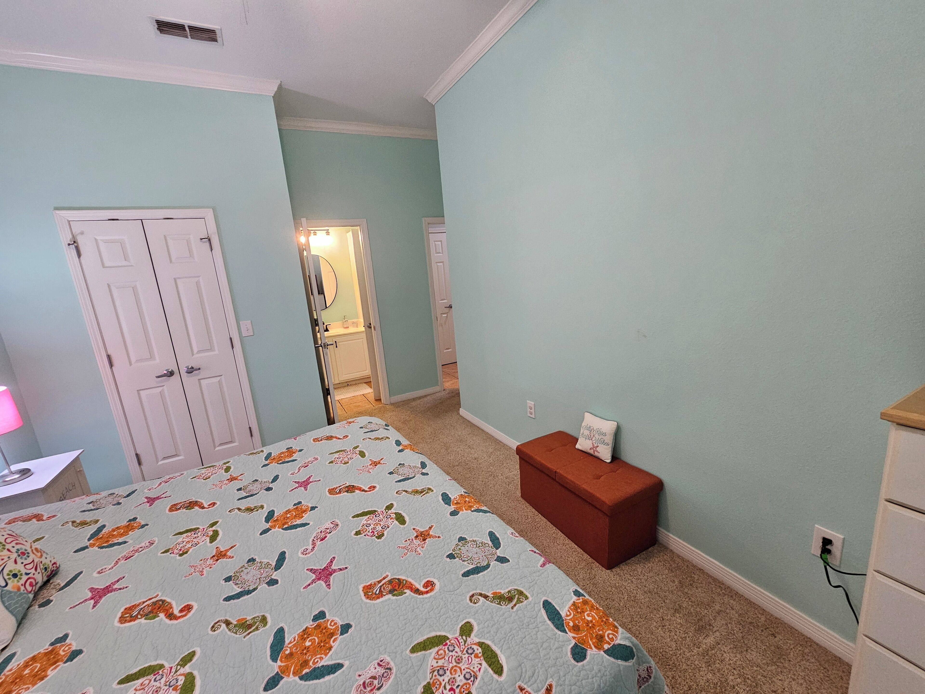 Une chambre à coucher comprenant un lit, une table de chevet, une commode et un petit banc. Une porte donne accès à la salle de bain.