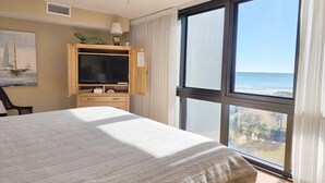 Una habitación de hotel con cama, televisión, ventana con vista a la playa y ventilador de techo.