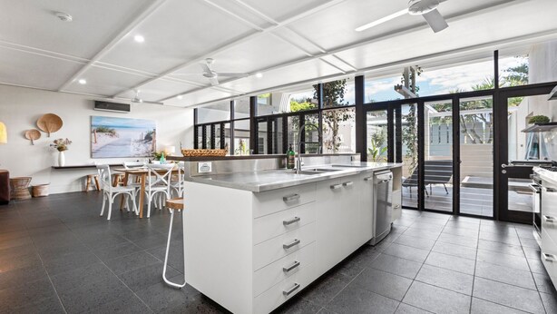 A modern kitchen with a large island, a dining area with white chairs, and a view of the outdoors through large windows. - Bronze @ Lennox Head (LENNOX HEAD)