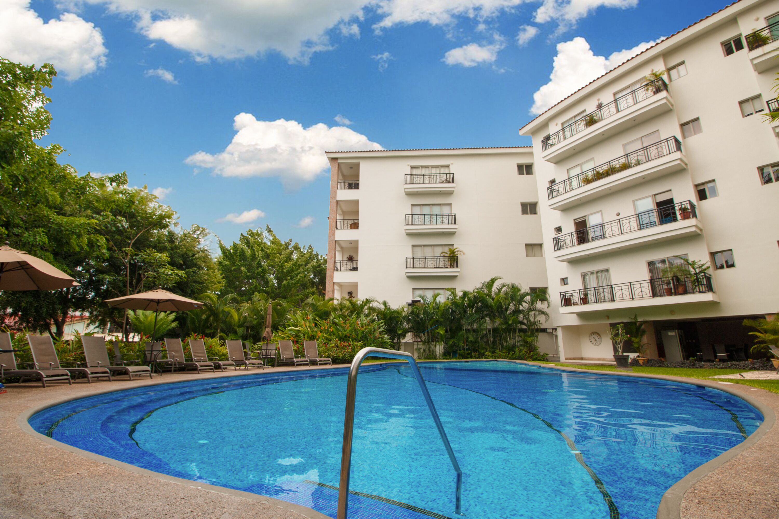 Piscina com escada de metal, cercada por espreguiçadeiras e guarda-sóis, com um edifício de vários andares ao fundo.