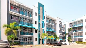 Un immeuble résidentiel moderne à plusieurs étages, doté de balcons, d’un espace de stationnement et de palmiers.