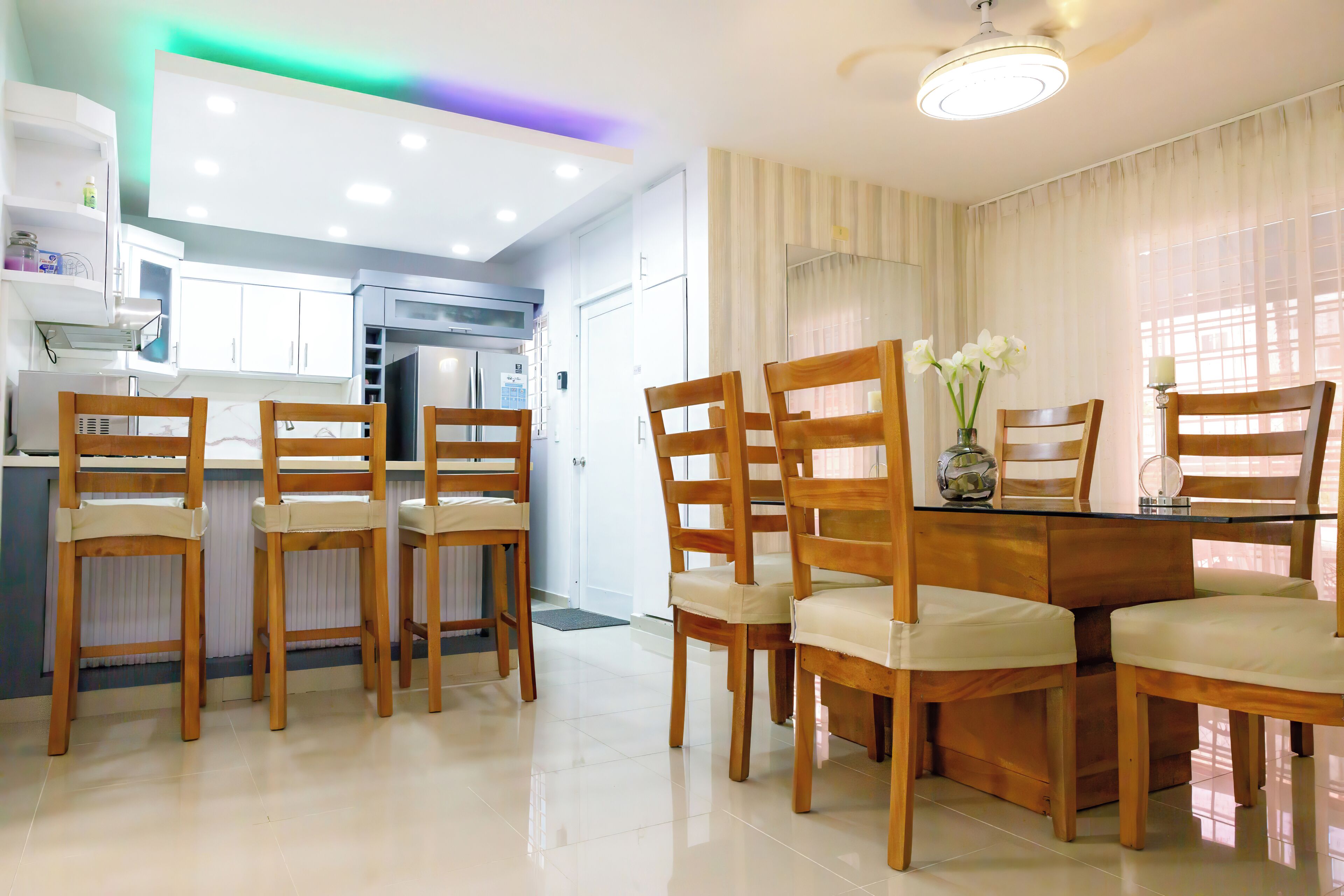 Une cuisine moderne avec des meubles blancs, une table à manger entourée de chaises en bois et un ventilateur de plafond.