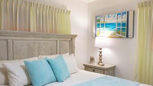Un lit bien fait, avec une tête de lit, une table de chevet sur laquelle se trouve une lampe et une petite statue, et un tableau représentant une scène de plage accroché au mur.