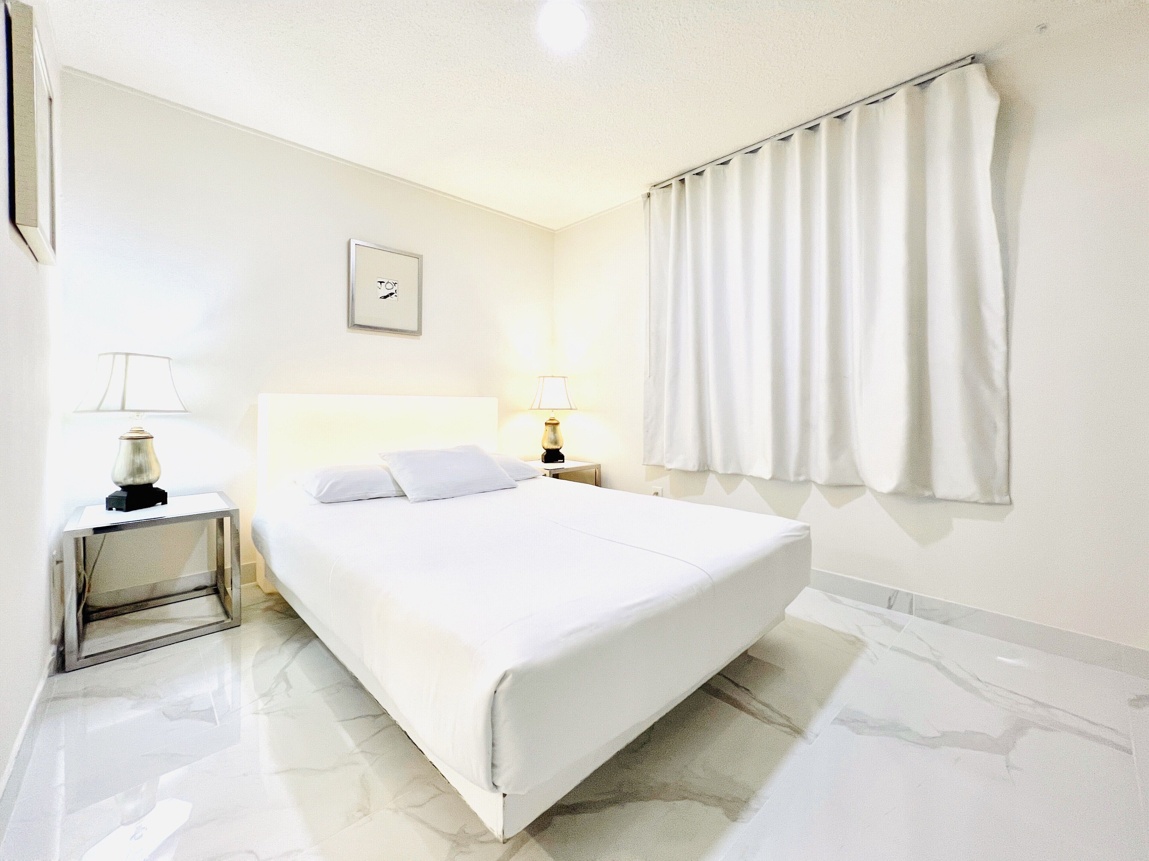 Une chambre d’hôtel avec un grand lit, deux tables de chevet, une commode, un tableau encadré au mur et une fenêtre avec des rideaux.