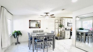 Een moderne keuken met witte kastjes, een eettafel met stoelen, een plafondventilator en een grote spiegel aan de muur.