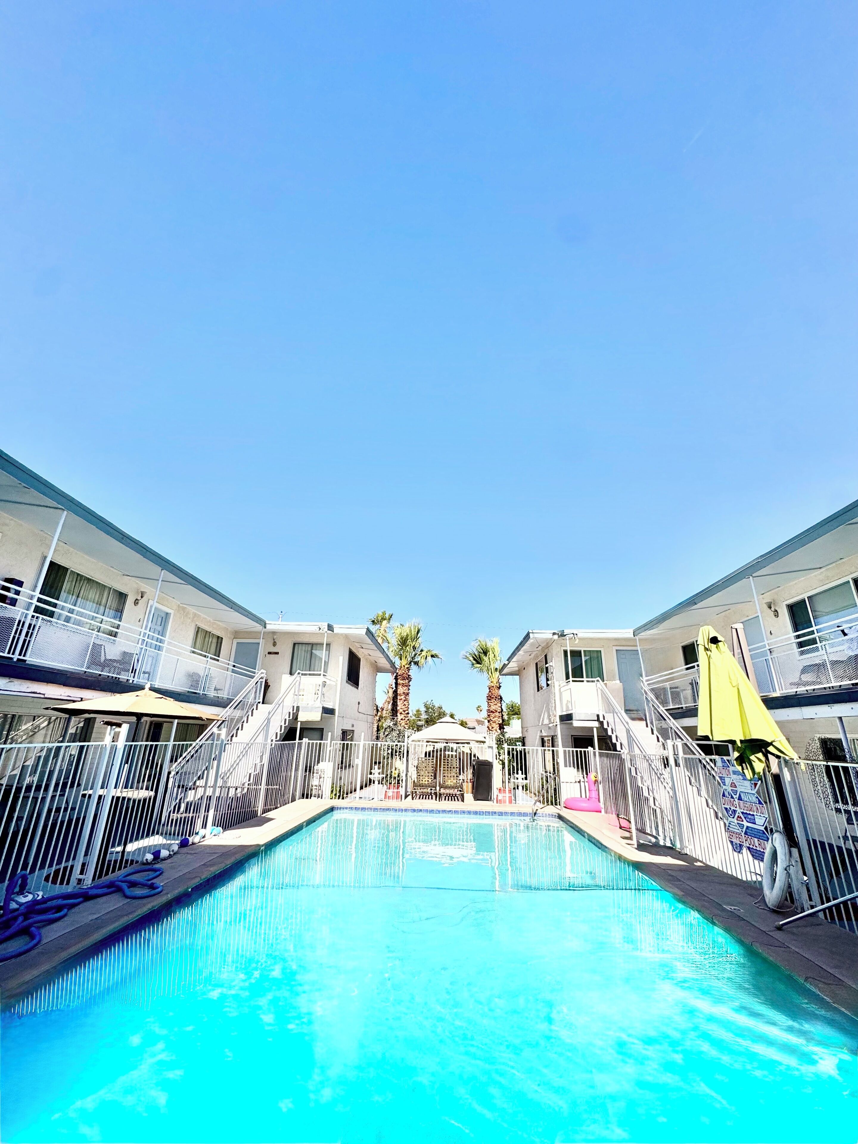 A swimming pool with a clear blue sky above, surrounded by a building with balconies and palm trees.