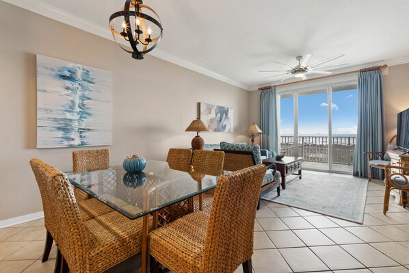 A spacious dining area with wicker chairs and a glass table, a ceiling fan, and a view through large windows.