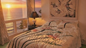 A bedroom with a bed, a nightstand, a lamp, a window with a view of the sunset over the ocean, and a painting on the wall. - SOUND FRONT / Kite-in/out Direct Beachfront Access Sunsets Best Kiteboard Spot (Rodanthe)