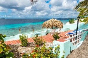 Un patio frente a la playa con un toldo de paja, sillas de descanso y vistas al océano.