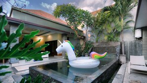 A swimming pool with a unicorn float, surrounded by greenery and a modern house. - Krishna Villas (Legian)