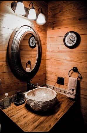 A rustic bathroom with a wooden vanity, a large round mirror, a stone sink, and a wall-mounted clock.
