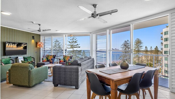 A modern living room with a dining area, large windows, and a ceiling fan.