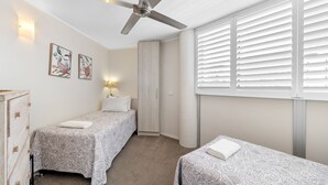A bedroom with two beds, a ceiling fan, a nightstand with a lamp, and a window with blinds.