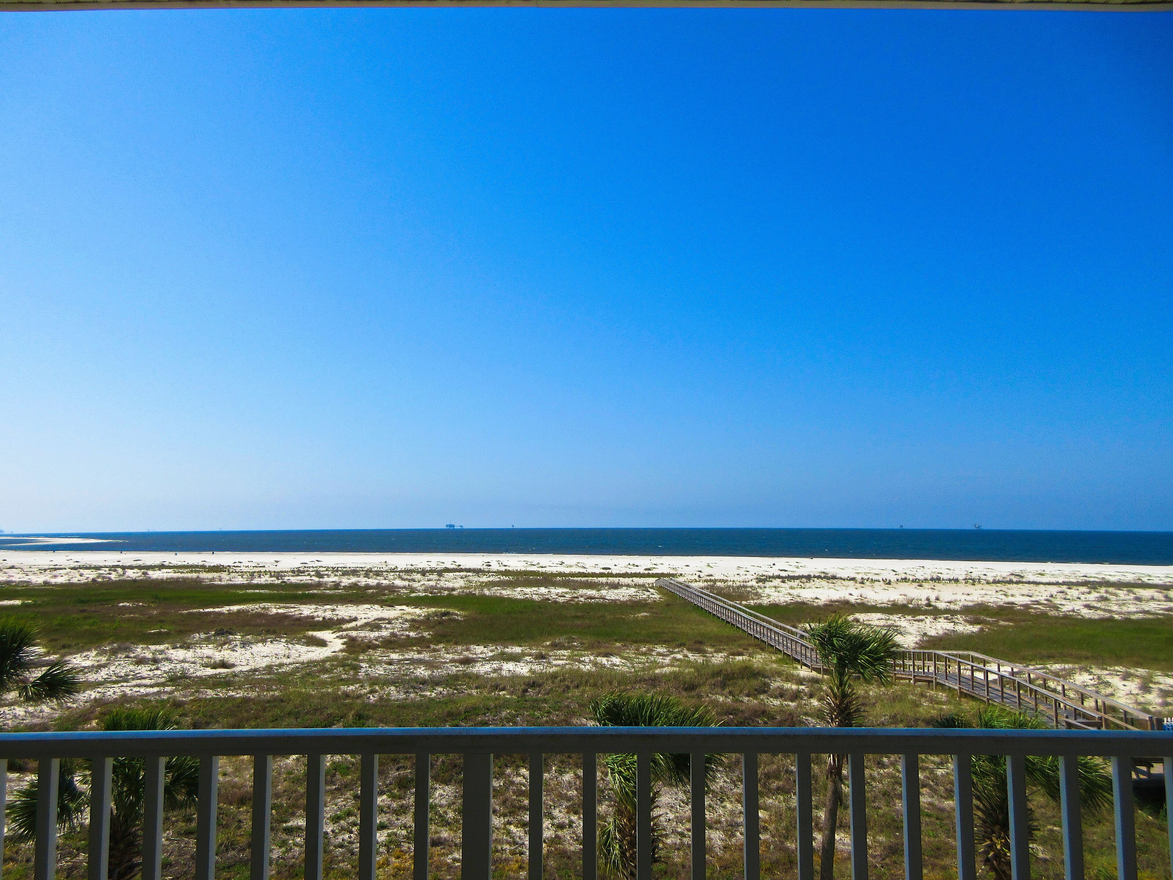 A beach view with a wooden boardwalk leading to the water, surrounded by sand and sparse vegetation.